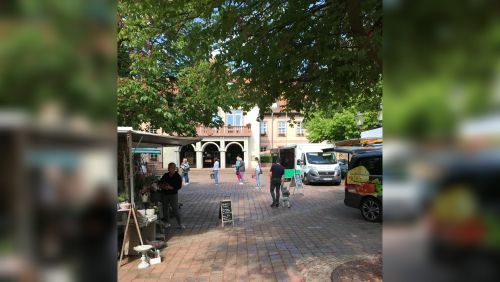 Gepflasterter Marktplatz mit Marktständen, Menschen und Lieferfahrzeugen unter großen Baumkronen.